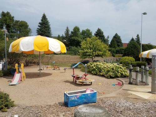 A playground with a large yellow and white umbrella, sand, slides, and play structures surrounded by greenery.