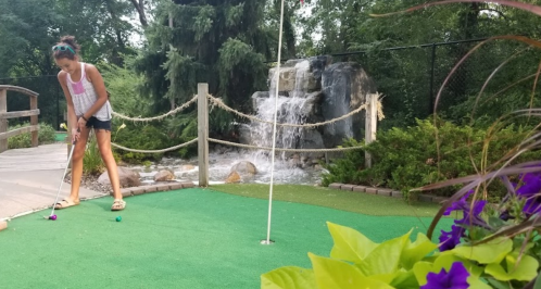 A girl plays mini-golf near a waterfall, surrounded by greenery and colorful flowers.