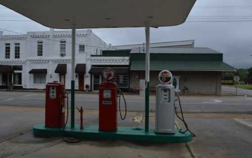 Three vintage gas pumps under a canopy, with a small town building in the background on a cloudy day.
