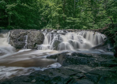 A serene waterfall cascades over rocks, surrounded by lush green trees in a tranquil forest setting.