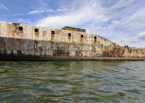 A weathered stone structure rises from the water under a blue sky, with patches of greenery and rust visible.
