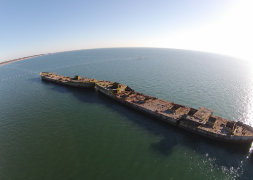 Aerial view of two rusted shipwrecks partially submerged in calm blue waters under a clear sky.