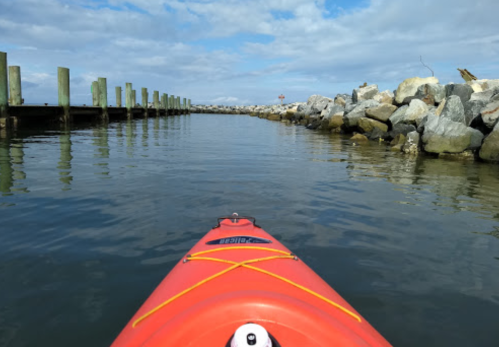 A red kayak on calm water, facing a rocky shoreline and wooden pier under a cloudy sky.