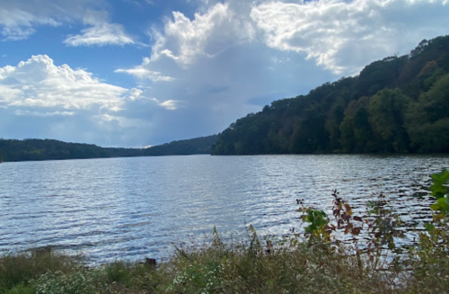 A serene lake surrounded by lush greenery and hills, under a partly cloudy sky.