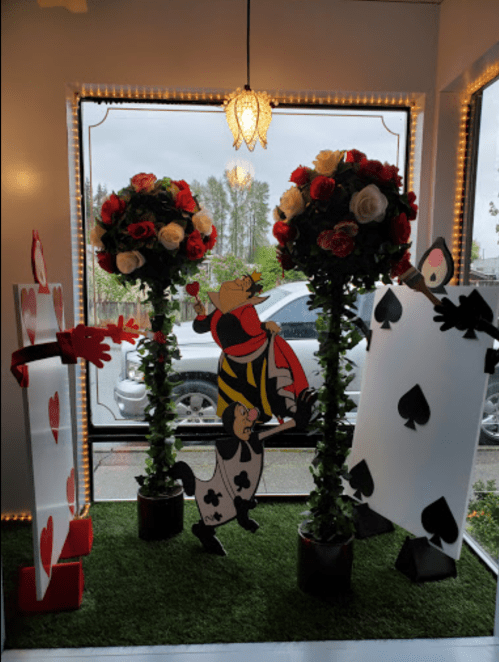 A whimsical display featuring large playing card cutouts, topiary roses, and a character in a striped outfit.