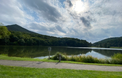 A serene lake surrounded by lush green hills under a cloudy sky, with a path along the water's edge.