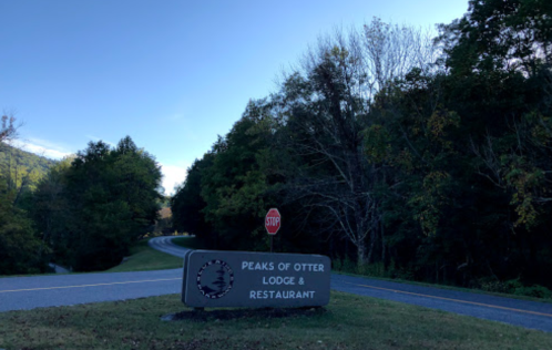 Sign for Peaks of Otter Lodge & Restaurant near a winding road, surrounded by trees under a clear blue sky.