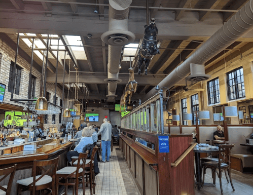 Interior of a restaurant with a bar, wooden seating, and large windows, featuring patrons and a modern industrial design.