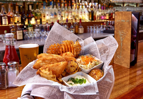 A basket of fried food with coleslaw, waffle fries, and grilled lemon, alongside a pint of beer at a bar.