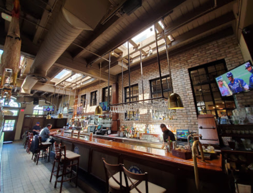 Interior of a bar with a wooden counter, brick walls, large windows, and a TV showing a sports game.