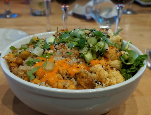A bowl of quinoa topped with roasted cauliflower, chickpeas, green onions, and a drizzle of sauce, garnished with herbs.