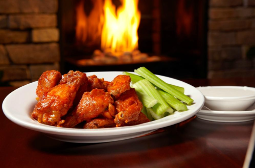 A plate of spicy chicken wings with celery sticks and dipping sauce, set against a cozy fireplace backdrop.