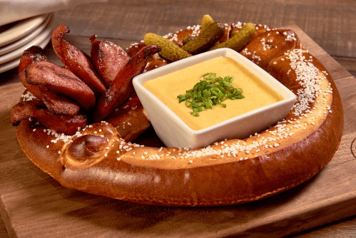 A large pretzel served with sliced sausage, pickles, and a bowl of cheese dip, garnished with green onions.