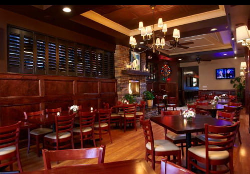 Cozy restaurant interior with wooden tables, chairs, and decorative lighting, featuring a stone fireplace and plants.