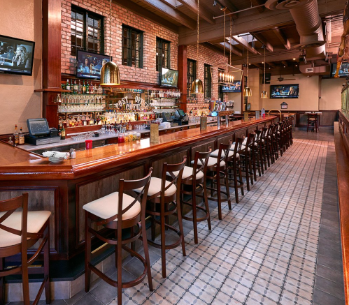 A spacious bar with wooden seating, brick walls, and multiple TVs displaying sports. The atmosphere is inviting and modern.