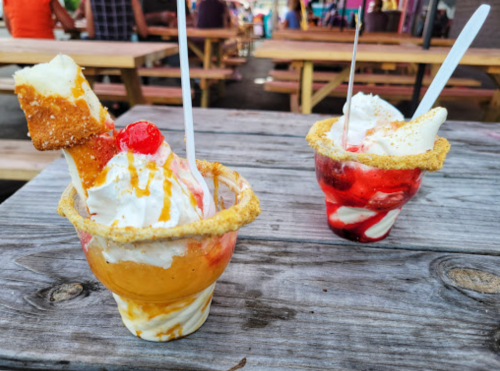 Two colorful ice cream desserts in cups, topped with whipped cream, cherries, and garnished with cookie rims, on a wooden table.