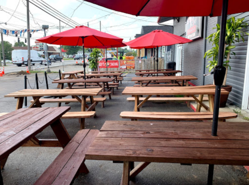 Outdoor seating area with wooden picnic tables and red umbrellas, set against a gray building and power lines.