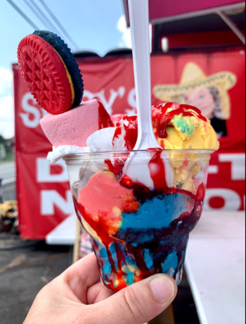A colorful ice cream sundae in a cup, topped with whipped cream, syrup, an Oreo, and a marshmallow.