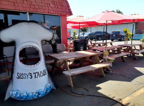 A whimsical taco stand with a shark-shaped entrance and picnic tables under red umbrellas.