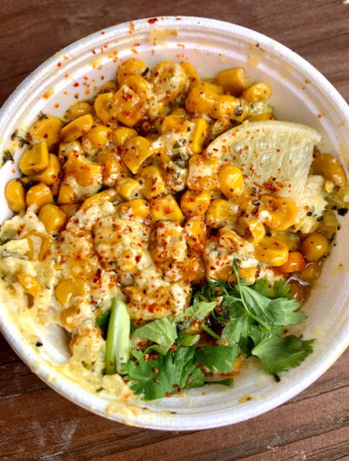 A bowl of corn topped with spices, cilantro, and a lemon wedge, served on a wooden surface.