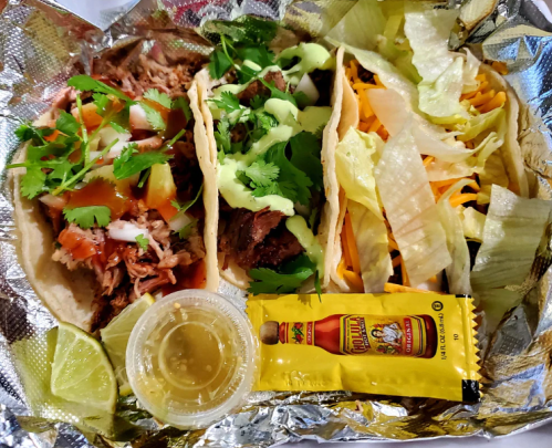 Three tacos filled with meat, topped with cilantro and sauce, served with lime and hot sauce on a foil tray.