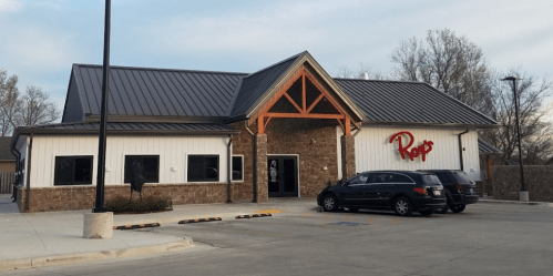 A modern building with a metal roof and stone accents, featuring a sign that reads "Ropo's" and parked cars in front.