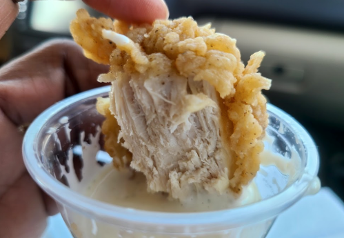 A hand holding a piece of fried chicken with a crispy coating, partially dipped in a creamy sauce.