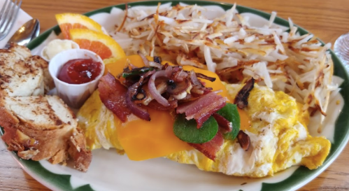 A plate of a cheesy omelet topped with bacon and vegetables, served with hash browns, toast, and a side of fruit.