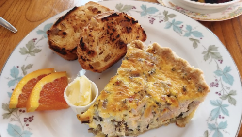 A plate with a slice of quiche, toasted bread, orange slices, and a small cup of butter.