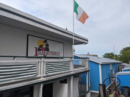 Sign for Idler Riverboat with an Irish flag flying above, set against a backdrop of blue buildings and a clear sky.
