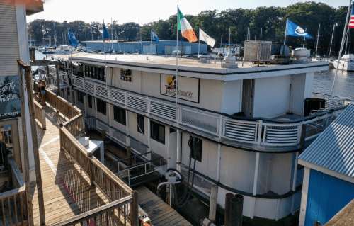 A large boat restaurant docked at a marina, featuring flags and a wooden walkway leading to it.