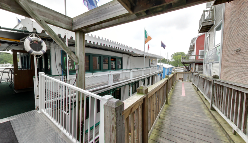A wooden walkway leads to a building with flags and an "Open" sign, surrounded by a waterfront setting.