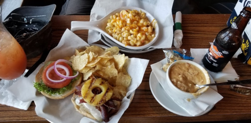 A table with a burger topped with bacon and pineapple, chips, mac and cheese, and a soda.