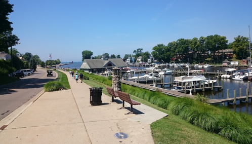A scenic waterfront view with a walkway, boats docked, and people enjoying a sunny day by the water.
