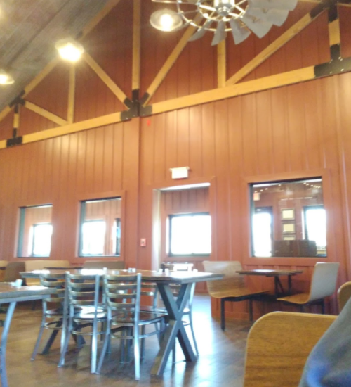 Interior of a rustic restaurant with wooden walls, large windows, and tables set for dining. Brightly lit and spacious.