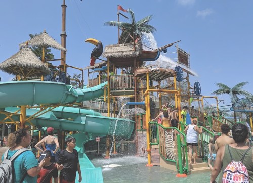 A vibrant water park with slides, a large play structure, and people enjoying the attractions under a clear blue sky.