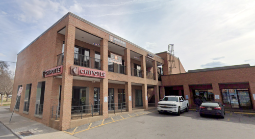 Exterior view of a Chipotle restaurant in a brick building, with a parking lot and nearby vehicles.