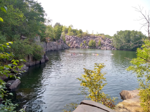 A serene lake surrounded by rocky cliffs and trees, with people swimming in the water on a sunny day.