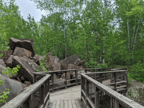 A wooden walkway leads through a lush green forest with large boulders scattered nearby.