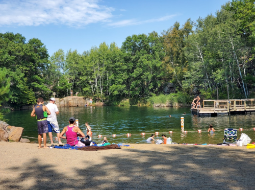 A sandy beach area by a calm lake, with people swimming, sunbathing, and enjoying the outdoors surrounded by trees.