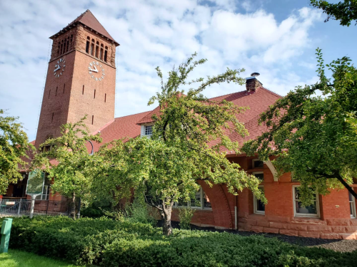 A brick building with a clock tower, surrounded by green trees and a blue sky with scattered clouds.