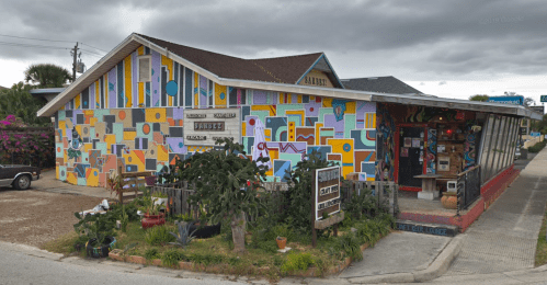 Colorful, abstract mural on a building with a garden and signs, set against a cloudy sky.