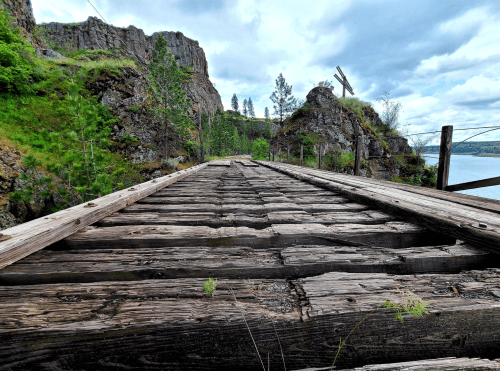 A weathered wooden bridge stretches over rocky terrain, surrounded by greenery and a calm body of water under a cloudy sky.
