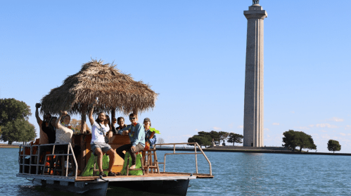 A group of people on a boat with a thatched roof, smiling and waving, near a tall monument by the water.