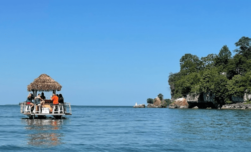 A group of people relax on a thatched-roof boat in calm waters, with lush greenery and cliffs in the background.