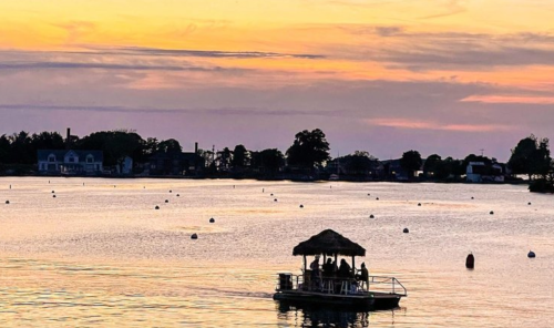 A boat with a thatched roof glides across a calm lake at sunset, surrounded by moored buoys and a colorful sky.