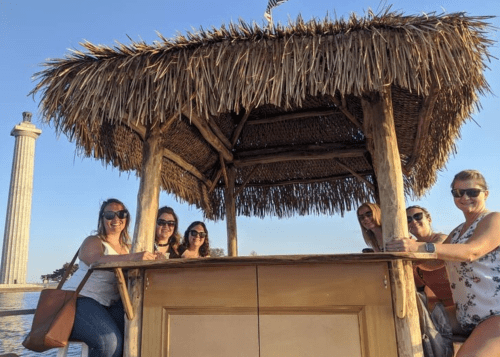 A group of six women smiles at a beachside bar with a thatched roof, enjoying a sunny day.