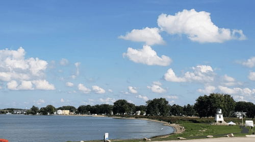 A serene lakeside view with fluffy clouds, green trees, and a small lighthouse in the distance.