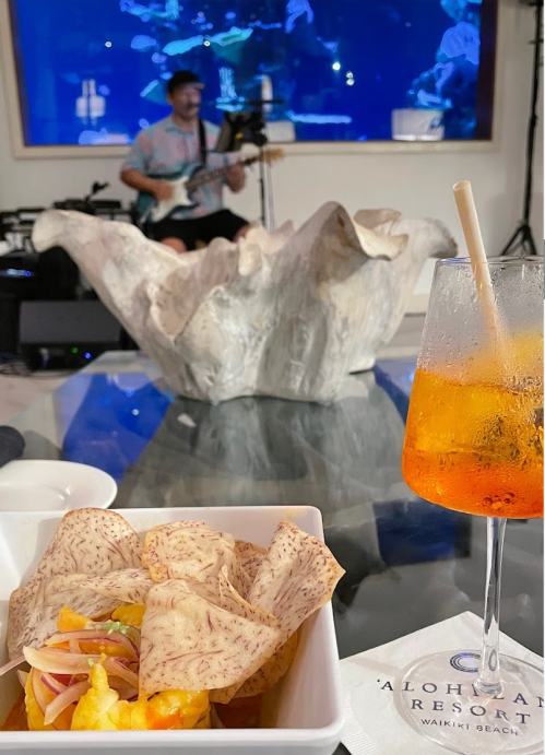 A vibrant cocktail and a dish of nachos in the foreground, with a musician playing in the background at a resort.