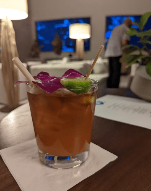 A cocktail garnished with purple flowers and lime, served in a glass on a wooden table with a blurred background.